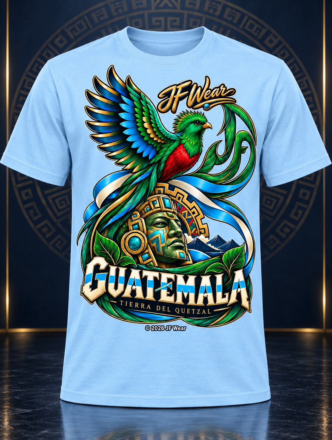 A light blue short-sleeve t-shirt featuring a large, colorful graphic print of a green and red quetzal bird, a golden mask, and the text 'I Wear GUATEMALA' and 'TIERRA DEL QUEXTAL'.