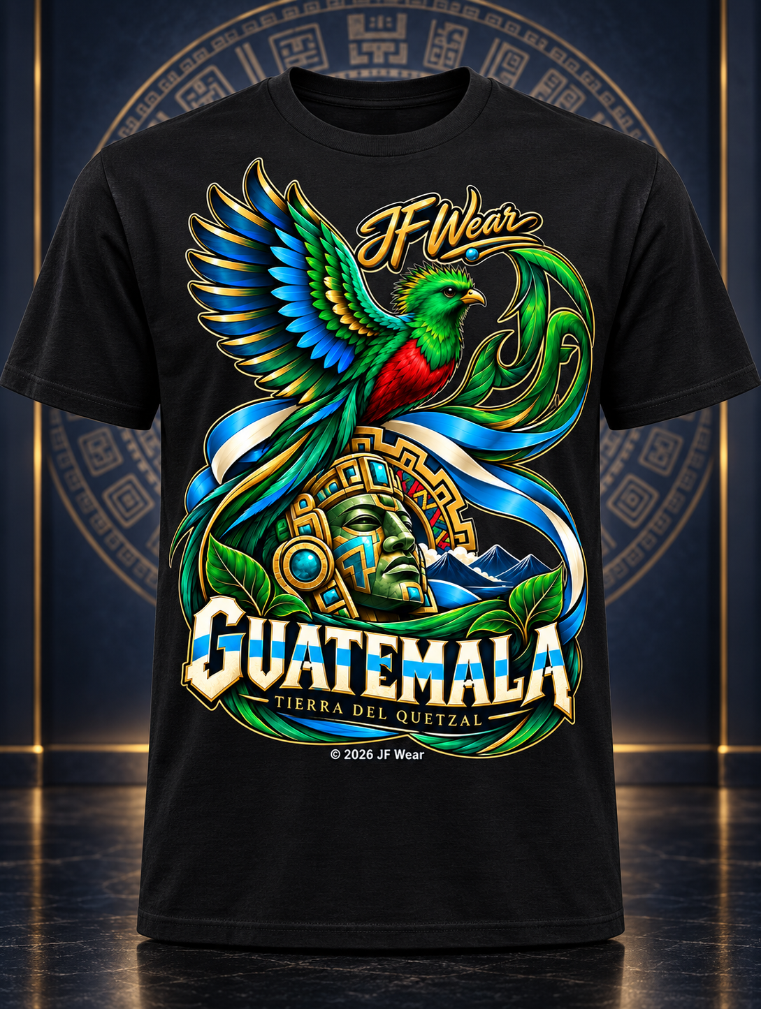 A black short-sleeve graphic T-shirt displayed front-facing against a dark blue and gold decorative backdrop. The shirt features a large colorful print with a green and red bird, a Mayan-style face, blue and gold scrollwork, and the text 'I Wear GUATEMALA' and 'TIERRA DEL QUINTAL.