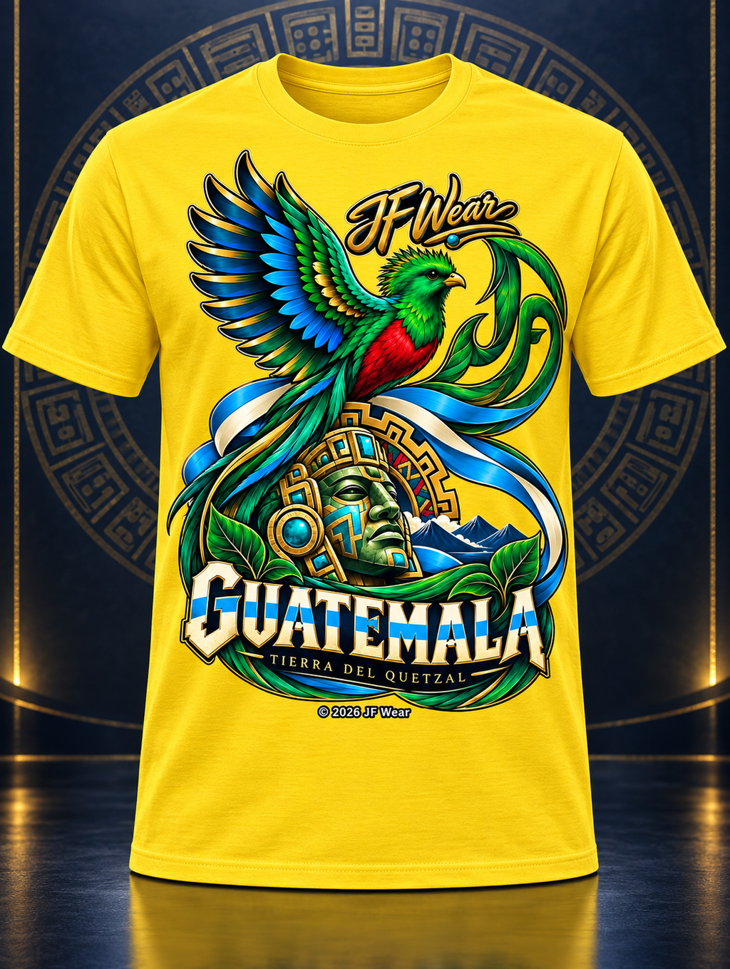 A bright yellow short-sleeve t-shirt featuring a large, colorful graphic print of a Guatemalan quetzal bird, a stone face, and the text 'IF Wear' and 'GUATEMALA' on the front, displayed against a dark blue background with a subtle circular pattern.